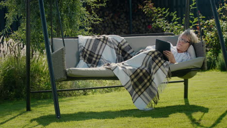 happy senior woman lies under a blanket in a garden swing, using a tablet. relaxing in the backyard of the house