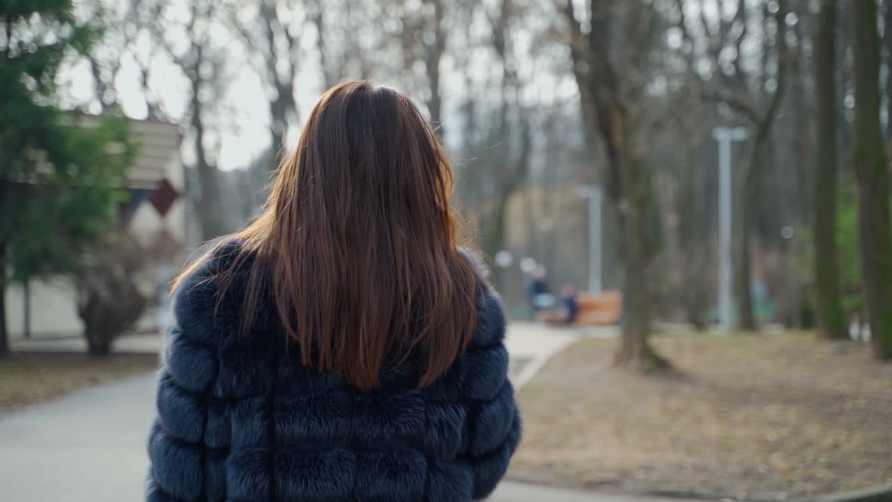 Woman in black luxury fur coat. Beautiful model with long dark hair walking in the park and drinks coffee. Back view. Slow motion.