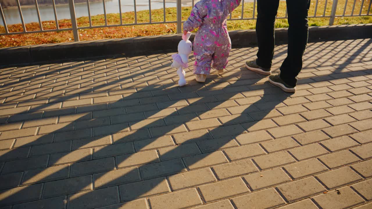 A child in a pink outfit walks with an adult, holding a toy, showcasing cozy companionship in the sunshine along a scenic, textured pathway
