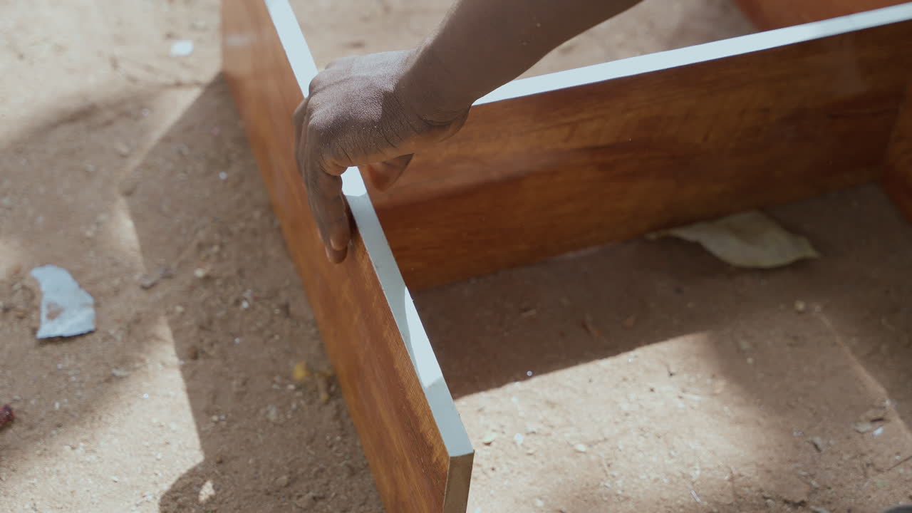 Primo piano di un falegname che assembla l'angolo di una cornice di legno in un cantiere polveroso, allineando il giunto a mano e controllando l'incastro sotto la luce del sole, una costruzione pratica di mobili che trasmette abilità costante e posizionamento misurato.
