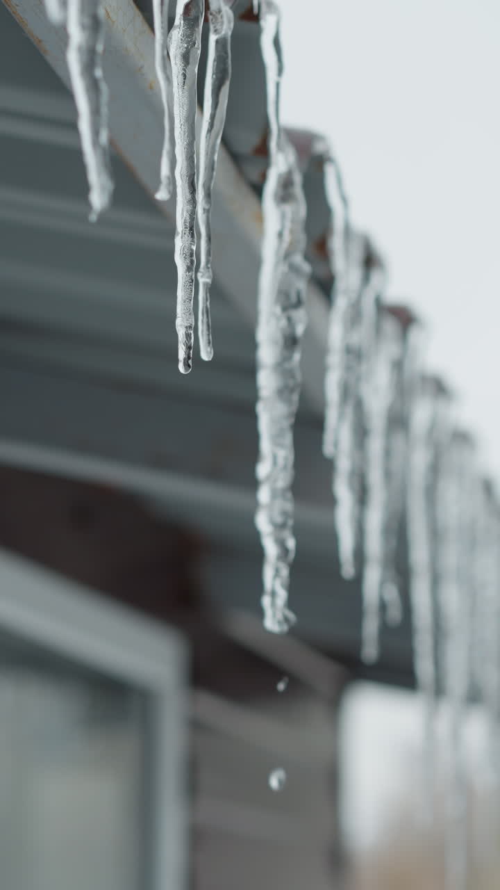 primer plano del borde del techo de la casa alineado con largos hielos que se derriten y gotean gotas de agua, mostrando intrincados detalles congelados y deshielo natural contra un fondo urbano suave y borroso durante el invierno