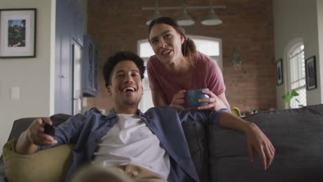 happy biracial couple sitting on sofa in living room, watching tv and drinking coffee