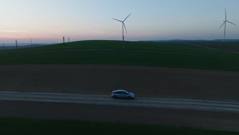 A-car-drives-on-a-rural-road-past-wind-turbines-at-sunset