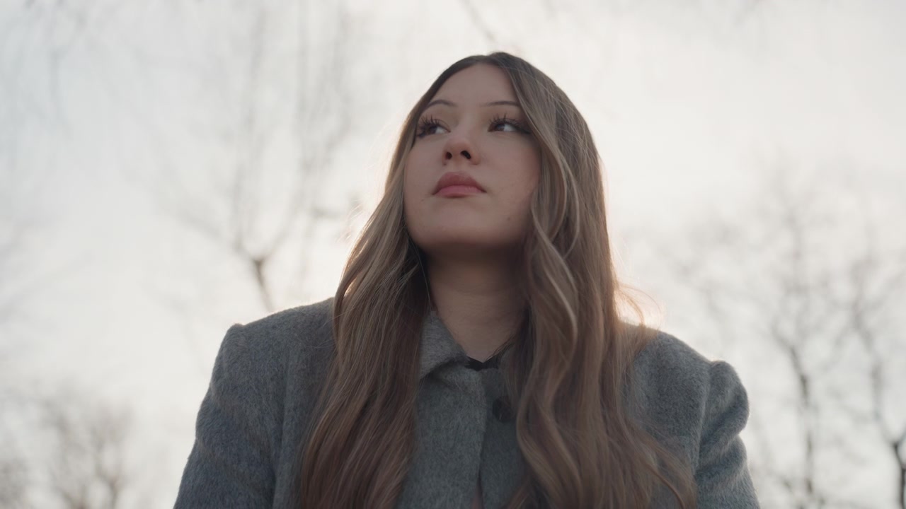Bella donna vicino ad alberi spogli, sorriso dolce, capelli lunghi, cappotto grigio, luce invernale nuvolosa, ritratto ravvicinato, umore introspettivo, leggera inclinazione della testa, profondità di campo cinematografica ridotta, bellezza naturale ed emotiva, studentessa