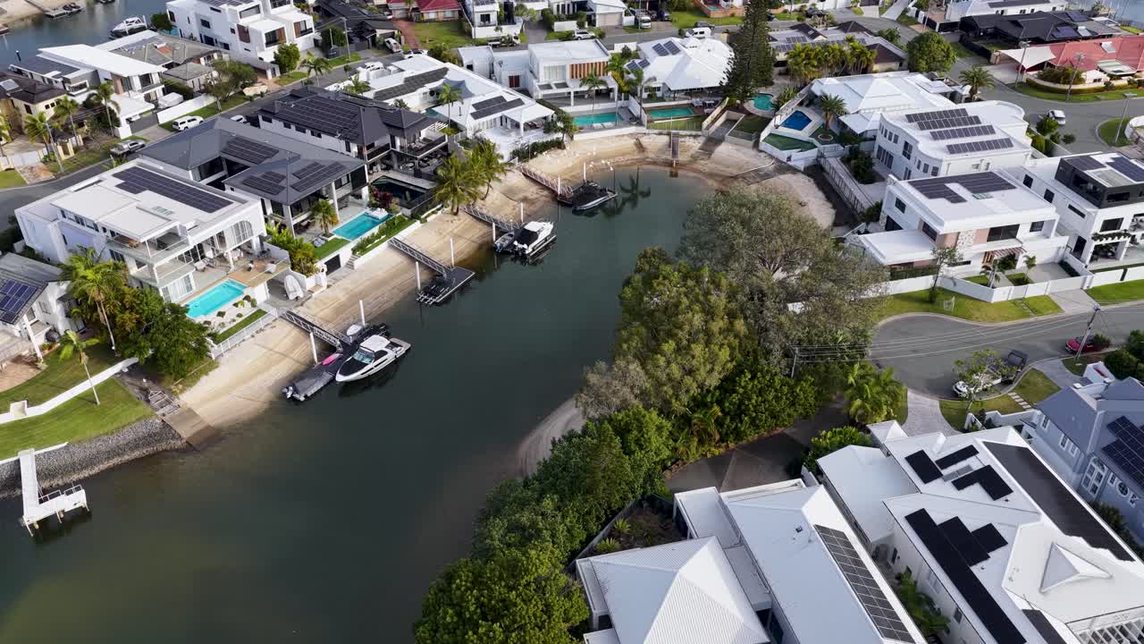 Drone camera glides above upscale riverside neighborhood with modern mansions, private pools, boats, and lush greenery in bright daylight, revealing waterfront lifestyle