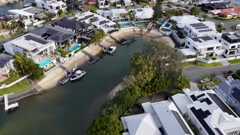 drone camera glides above upscale riverside neighborhood with modern mansions, private pools, boats, and lush greenery in bright daylight, revealing waterfront lifestyle