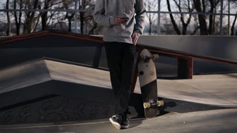 Young-Curly-Headed-Male-Skateboarder-Browsing-In-His-Mobile-Phone-In-The-Skate-Park-On-A-Sunny-Day