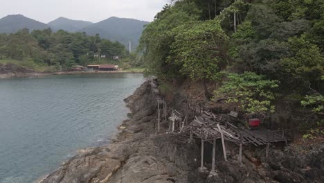 Drone-left-trucking-shot-of-a-wooden-deck-and-walkway-that-is-collapsing-onto-granite-rock-coastline-on-a-tropical-Island-with-jungle-lush-green-forest-and-ocean