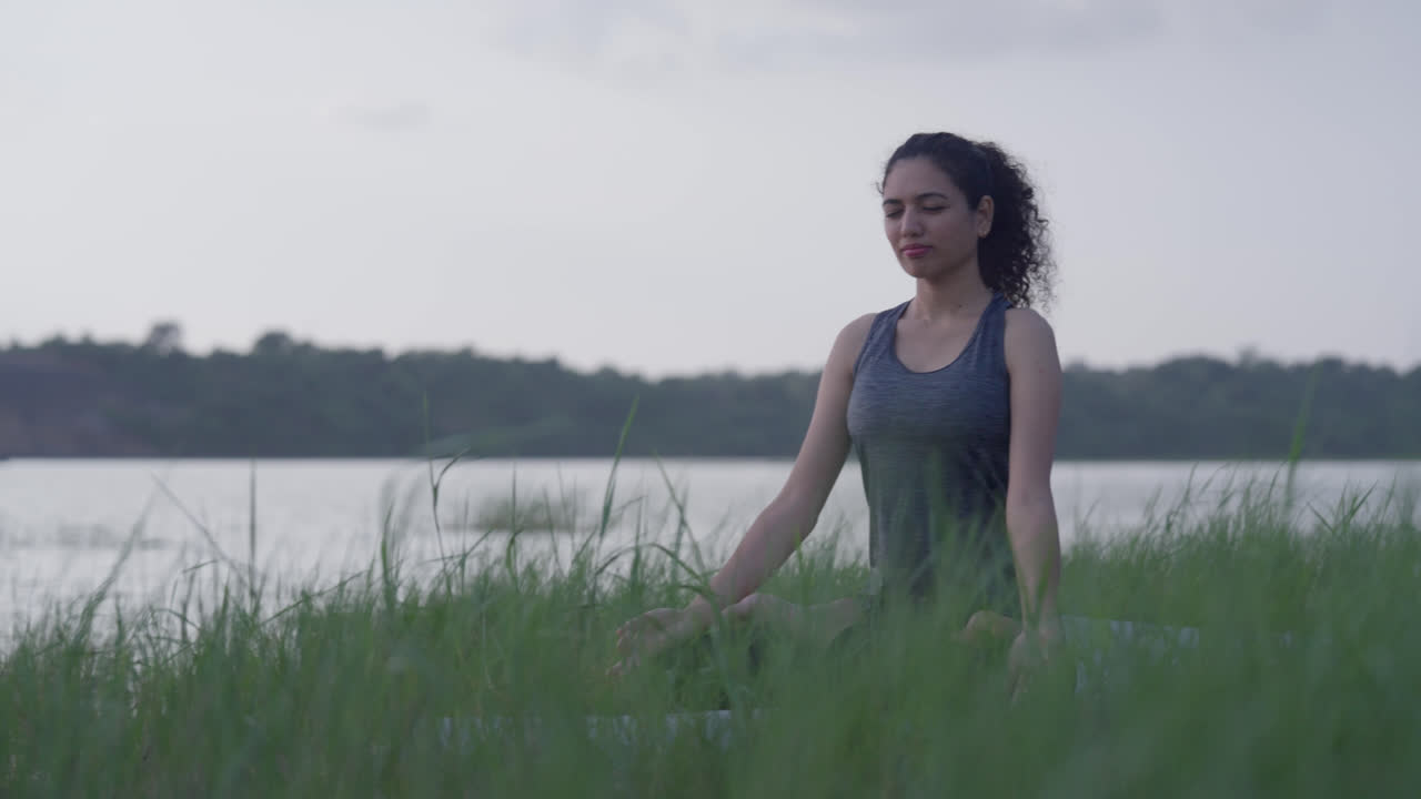 A South Asian Woman meditating in Lotus Pose (Padmasana) by a lake in tall grass during sunsetA South Asian Woman meditating in Lotus Pose (Padmasana) by a lake in tall grass during sunset