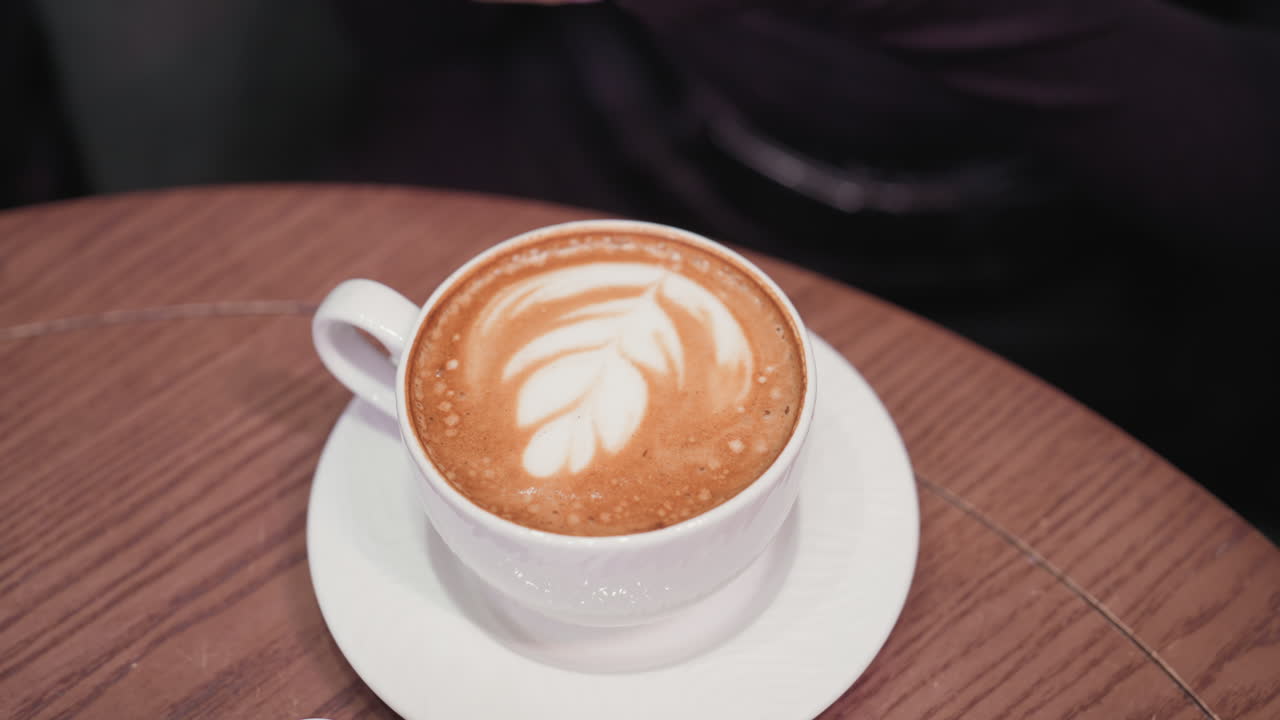 Close-up view of latte with elegant leaf-shaped foam art in white ceramic cup and saucer, placed on brown wooden table. Female hand holds smartphone nearby, capturing cozy coffee moment