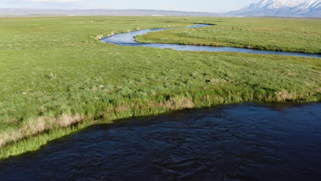 A-drone-flies-low-and-fast-over-a-beautiful-river-that-winds-its-way-through-green-fields-and-past-snow-covered-mountains-in-the-distance