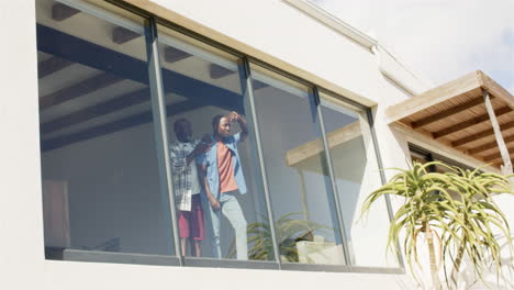 looking out window, african american father and adult son enjoying view from modern home interior