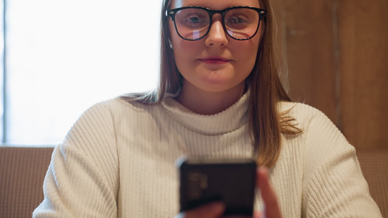 Young lady in white ribbed sweater smiles softly while using smartphone, with phone screen reflected in glasses, seated in cozy room as sunlight streams through window creating warm natural backlight