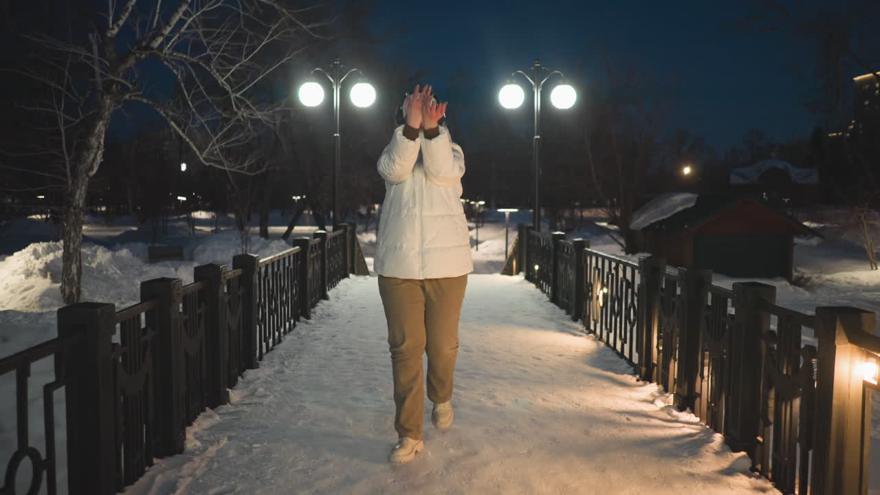 Girl wearing white puffer coat and headphones moves body in rhythm of music on illuminated snowy walkway near glowing lampposts and string lights with expressive gestures under silent winter night