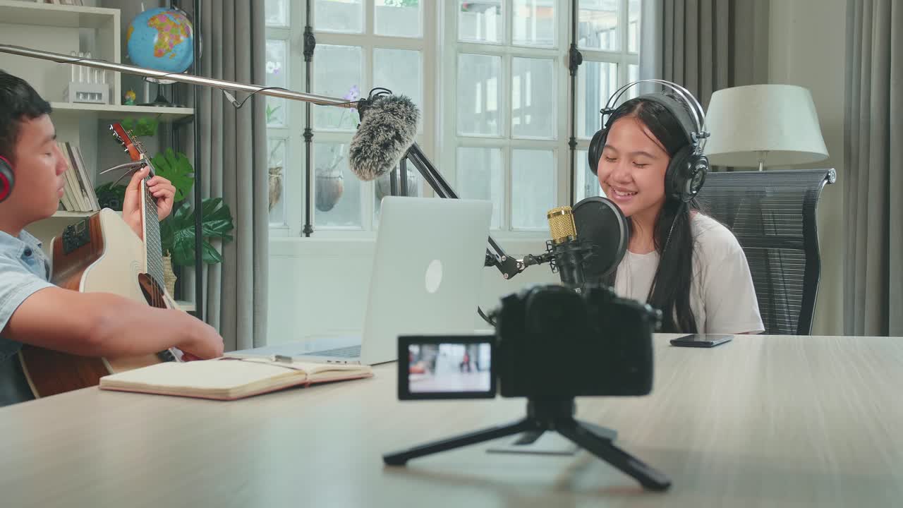 Camera Recording, Asian Kid Girl In Headphones Singing Into Microphone While Boy Playing The Guitar During Online Livestream In Podcast Studio