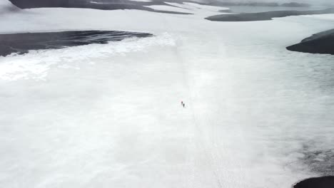 Gorgeous aerial footage flying closer and over two hikers walking on Iceland landscapes between mountains and frozen lakes during a cloudy day. The lakes have dark ice formed on them with cracks.