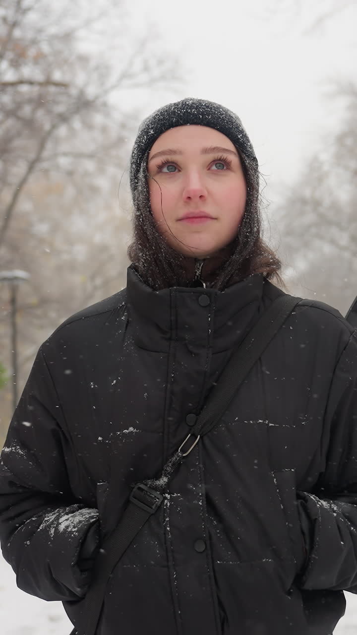검은 후디를 입은 여자가 주머니에 손을 고 기타를 들고, 눈이 내리는 눈길에 눈으로 인 길에서, 벌거벗은 나무와 조용한 겨울 풍경으로 둘러싸인 눈이 내린 길에서 사려 깊게 서 있습니다.
