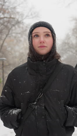 lady in black hoodie carrying guitar in pack with hands in pockets, standing thoughtfully in snowfall on a snow-covered path, surrounded by bare trees and serene winter scenery