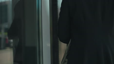 close up of professional woman with handbag opening glass door to enter building, focus on hand gripping metal handle, reflections on door surface with urban office environment