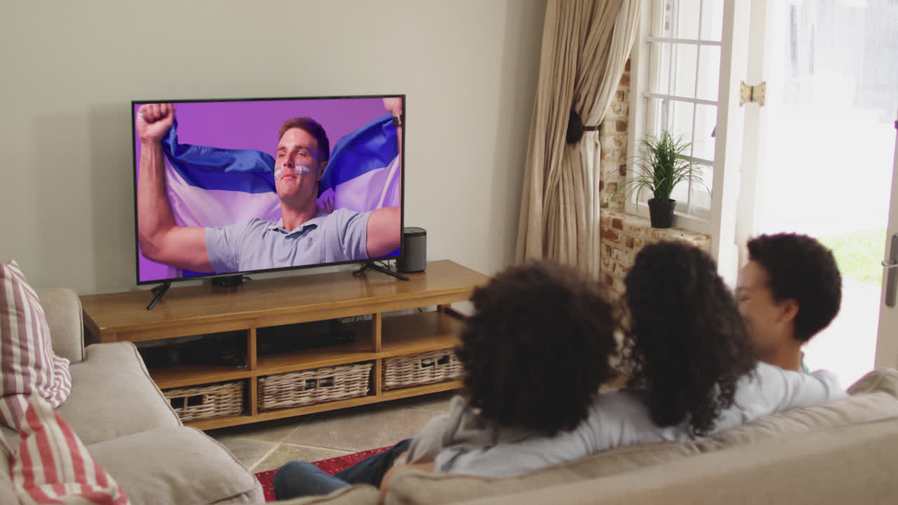 Famiglia birazziale che guarda la TV con un tifoso maschio caucasico con una bandiera sullo schermo