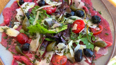 Delicious-traditional-gourmet-beef-carpaccio-with-parmesan-cheese,-cherry-tomatoes,-salad,-capres,-black-olives-and-pesto-sauce-on-a-plate-at-a-French-Italian-restaurant,-4K-shot