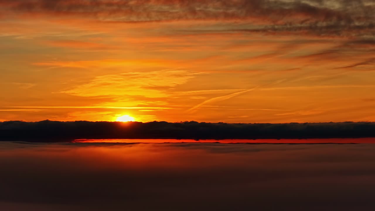 Eine leuchtend orangefarbene Sonne geht hinter einem dunklen Wolkenband unter und malt den Himmel mit warmen Tönen und sanften Spiegelungen über den Horizont
