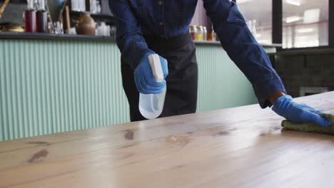 Mujer-Afroamericana-Con-Guantes-Y-Delantal-Desinfectando-Mesas-En-El-Bar-Cafetería