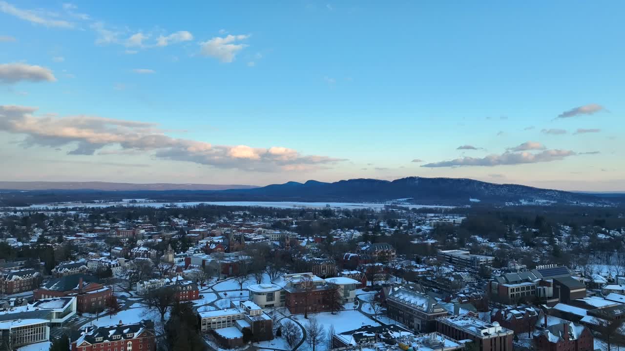 Drone footage of Northampton during winter, featuring snowy rooftops, historic buildings, and a scenic backdrop of mountains under a clear blue sky