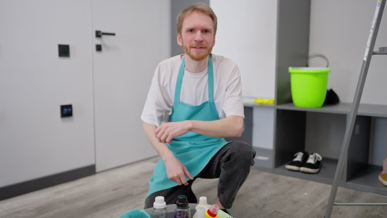 retrato en primer plano de un tipo confiado un limpiador rubio en una camiseta blanca y un delantal azul que puso detergentes y herramientas de limpieza en un lavabo de plástico gris mientras limpiaba en un apartamento moderno