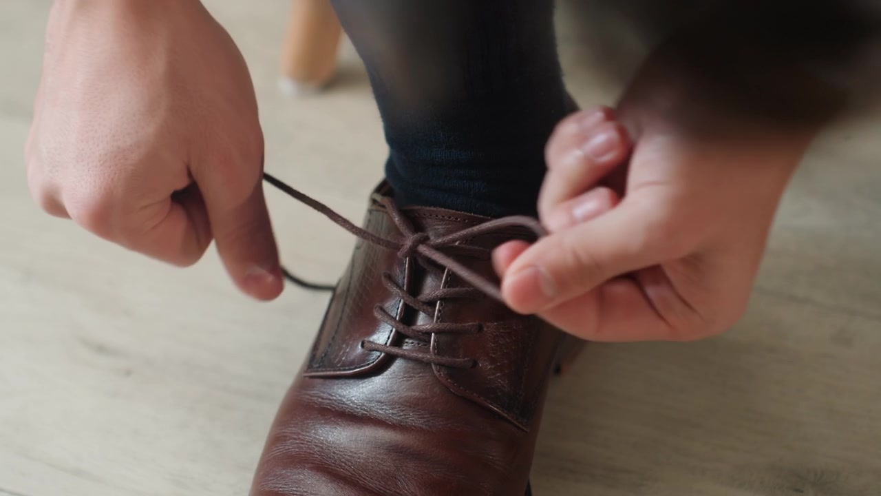 Uomo che allaccia una scarpa di pelle marrone, primo piano delle mani che annodano i lacci durante la calma preparazione mattutina per un matrimonio e un evento formale, pavimento in legno, calzino blu navy, pelle lucida, rituale concentrato e delicato.