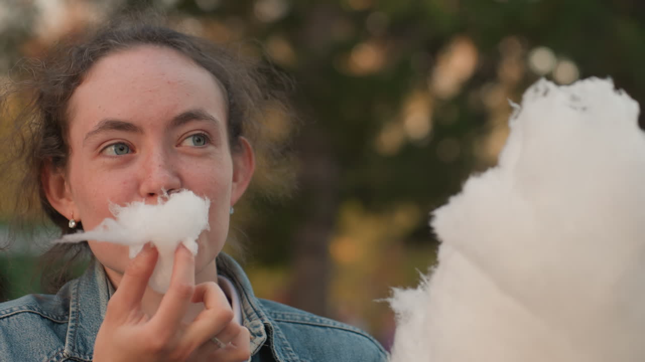 Donna sorride alla fiera notturna. Giovane donna assapora con gioia lo zucchero filato. Giovane appassionata si gode la soffice confezione di zucchero in un'atmosfera vivace di fiera e illuminazione brillante.