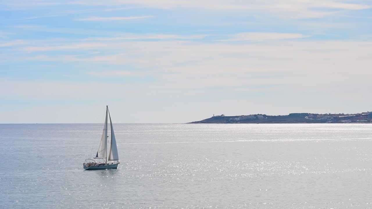 A single sailboat glides across the calm, shimmering Mediterranean Sea off the coast of Alicante, Spain. A peaceful and tranquil scene of leisure, travel, and the coastal lifestyle