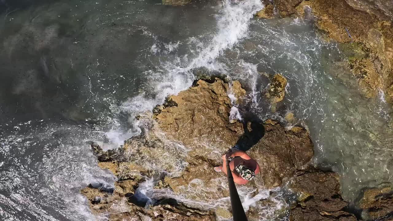High angle overhead view of man wearing hat walking on sea rocks holding long camera pole and circling