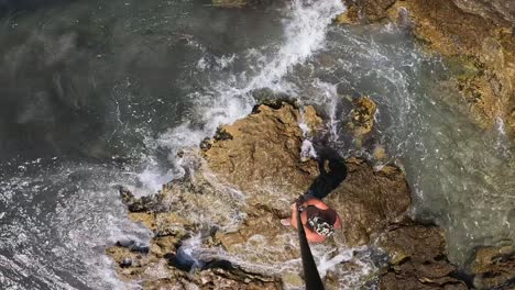 vista aérea en ángulo alto de un hombre con sombrero caminando sobre rocas marinas sosteniendo un largo poste de cámara y dando vueltas