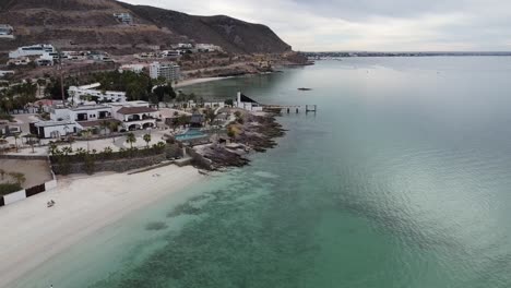 Ascending-aerial-shot-of-the-wonderful-beach-of-Playa-El-Caymancito-near-La-paz-Baja-California-Sur-Mexico-overlooking-the-turquoise-sea-and-the-beautiful-buildings-during-a-great-vacation