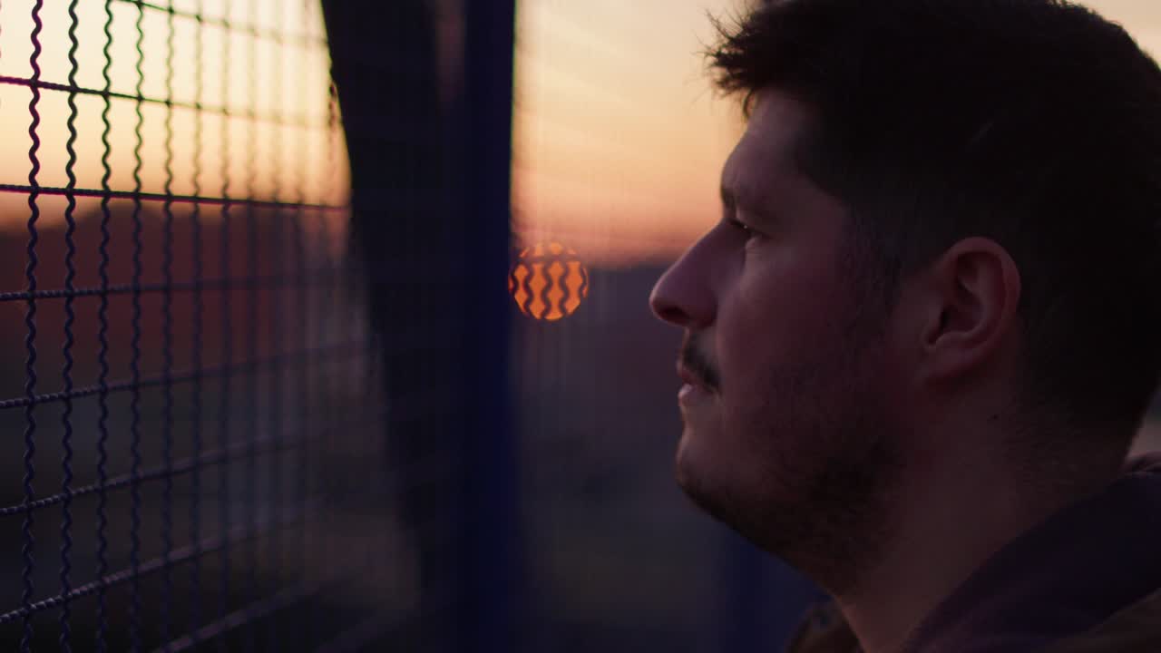 un hombre mira hacia la distancia a través de una valla de metal azul en un puente de hierro, su expresión triste visible. el cielo del anochecer sirve como un telón de fondo de mal humor, evocando una sensación de introspección y soledad.