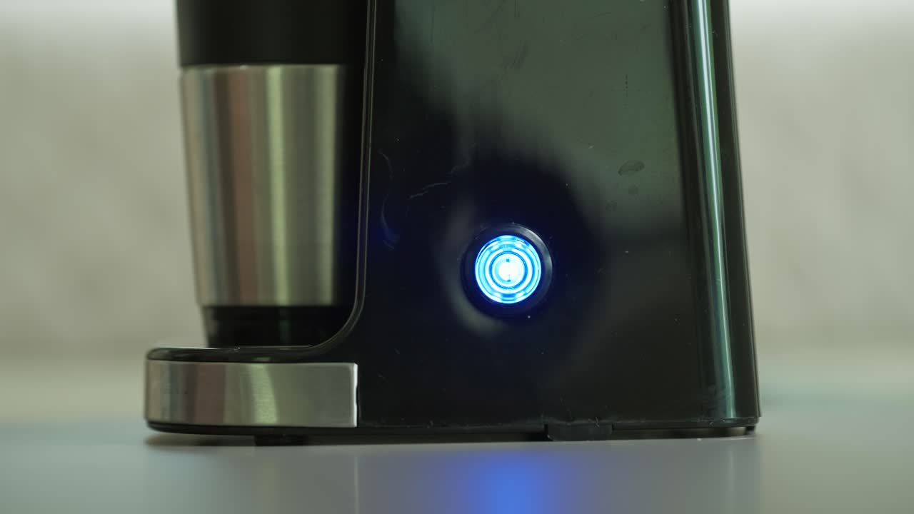 Una mano accende un pulsante con spia luminosa per avviare la preparazione del caffè in una piccola macchina da caffè americano.