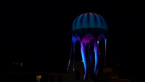 blue jellyfish balloon flying in the dark sky at night over the houses while it is being illuminated