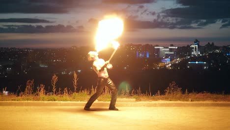 Young-blond-male-does-tricks-with-fire-breaths-fire-in-the-middle-of-night-with-city-skyline-background