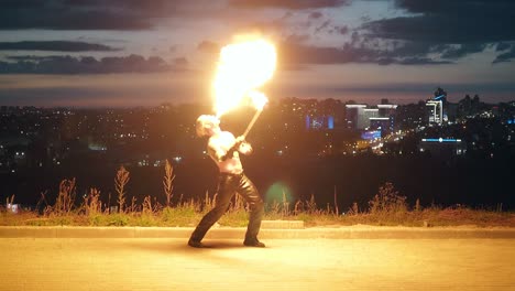 young blond male does tricks with fire breaths fire in the middle of night with city skyline background