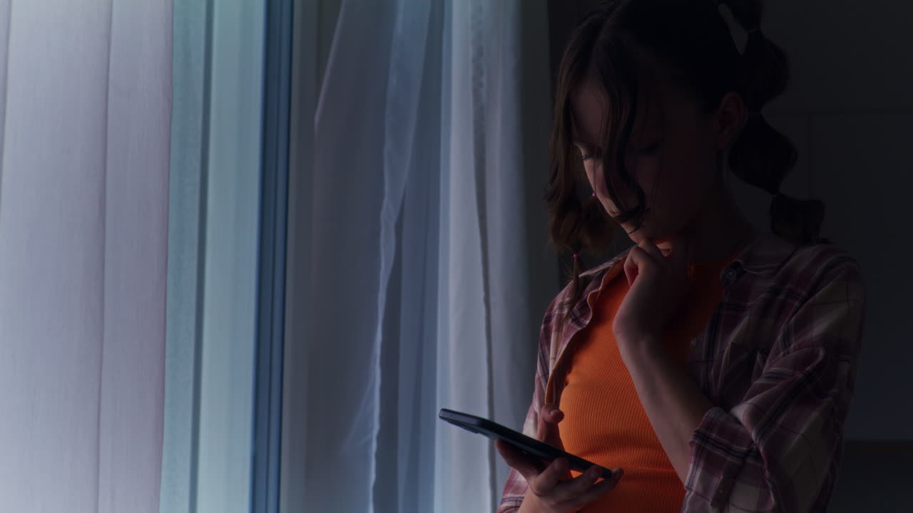 소셜 미디어에 중독된 어린 소녀가 창문 에 서서 스마트폰과 인터넷을 탐색합니다.