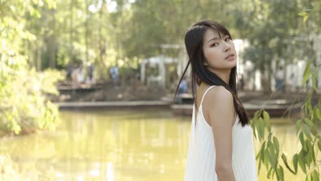 an asian woman in a white dress stands at the edge of a pond and looks at the camera