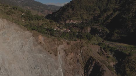 Aldea-Al-Borde-De-Un-Acantilado-En-Una-Región-Montañosa-Valle-Rodeado-De-árboles-Inversión-Aérea-Revelando-Una-Enorme-Pared-Rocosa-Kabayan-Benguet-Filipinas