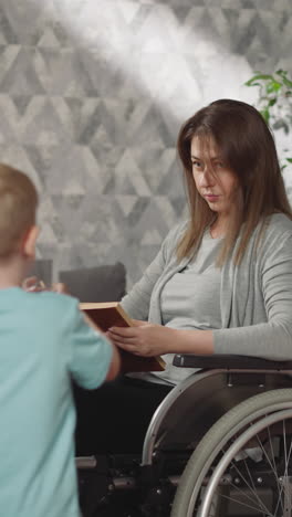 blond toddler brings glass of water to mom with disability. woman sits in wheelchair reading book while caring little boy helps mummy after surgery