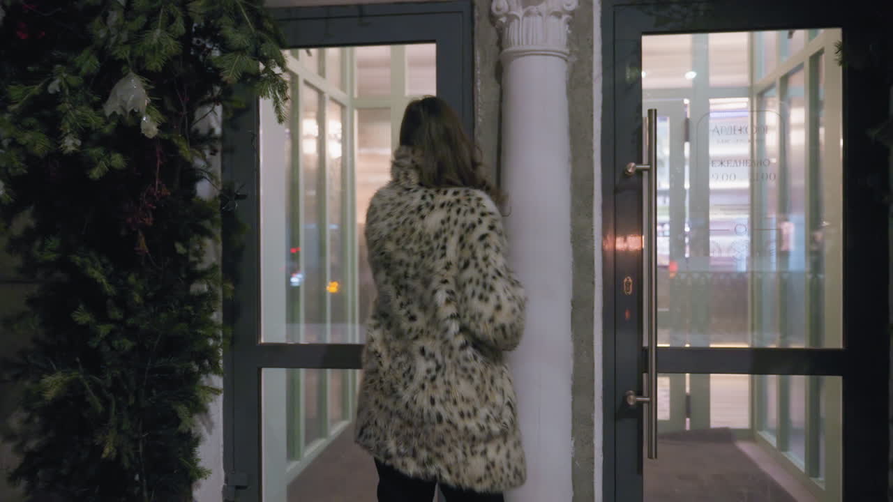 Woman with curly hair wearing spotted winter coat walks toward glass door entrance surrounded by snowy ground and festive greenery as door automatically closes behind her in cozy winter city setting