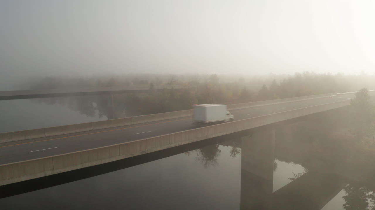 Commercial transportation, tractor trailer and box truck crossing highway bridge with blanket of fog hanging over river and landscape