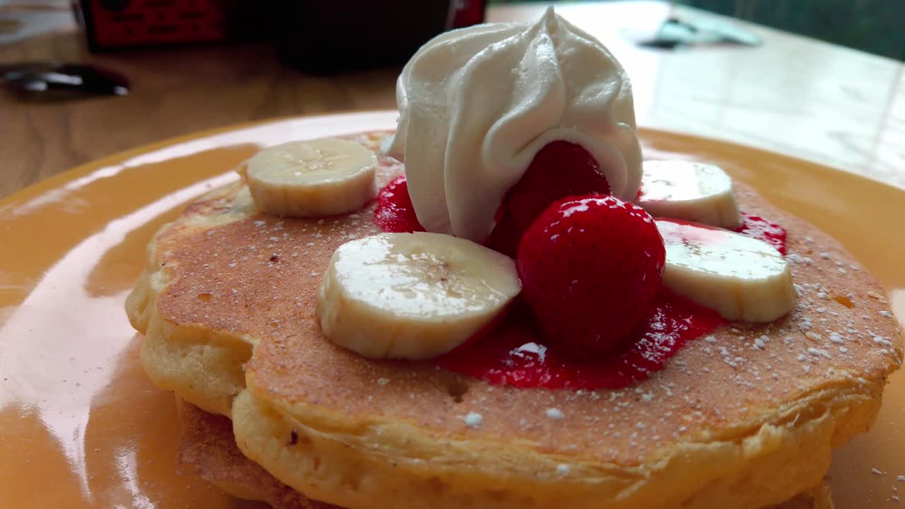Zoom out from fluffy pancakes with whipped cream, banana slices and strawberries served on a plate.