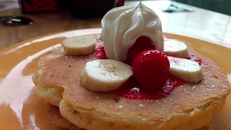 zoom out from fluffy pancakes with whipped cream, banana slices and strawberries served on a plate.