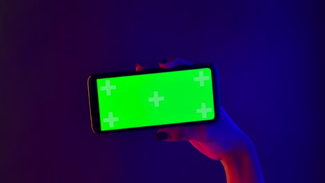 african american woman holds in her hand a smartphone with a green screen chroma key. close up of a phone in a horizontal position against a background of bright multicolored neon lights. slow motion