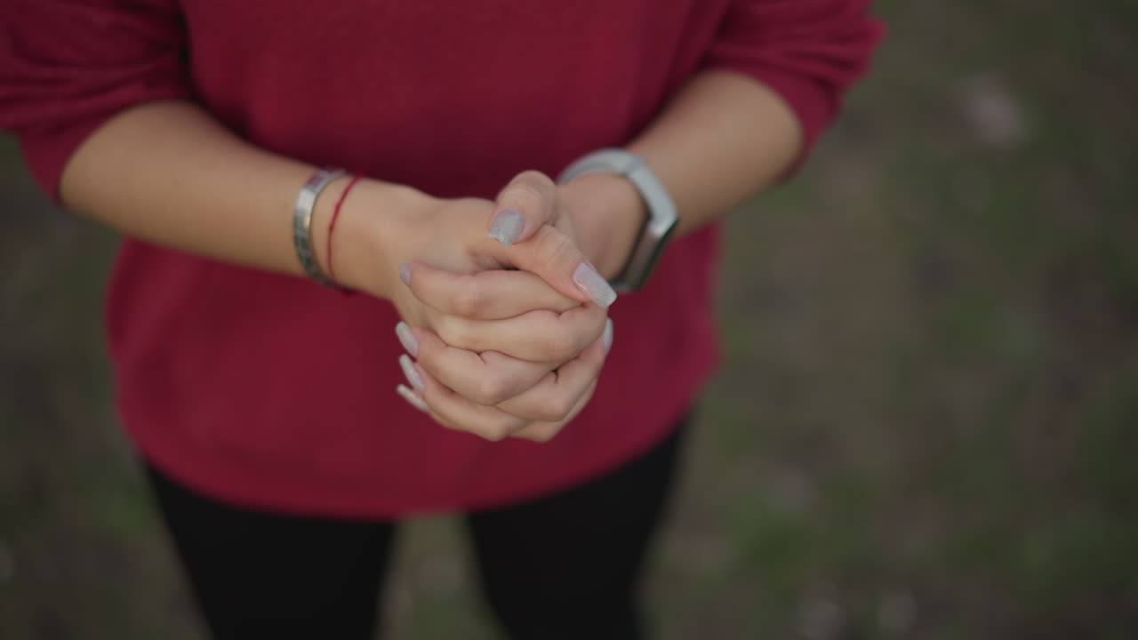 CloseUp Hands Warming Before Run Outdoors. Red Hoodie, Smartwatch And Bracelets Visible, Manicured Nails, Gentle Rubbing Motion To Loosen Fingers, Soft Grassy Park Background, Moody Light And Calm